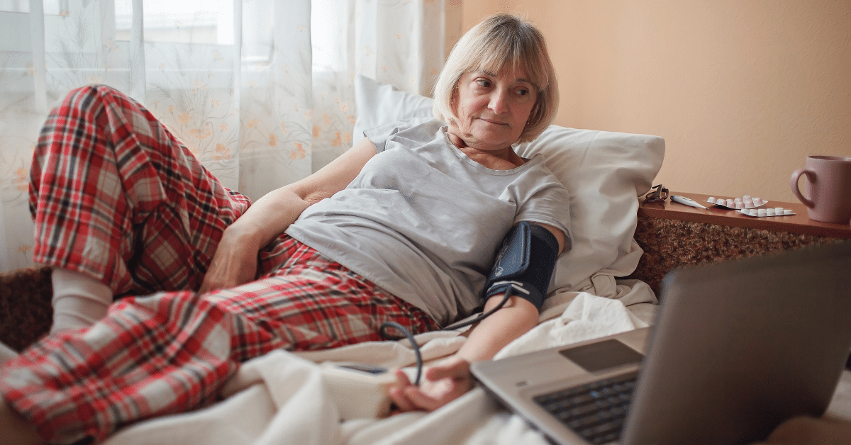 Woman in pajamas lying in bed looking at laptop while taking blood pressure next to medication